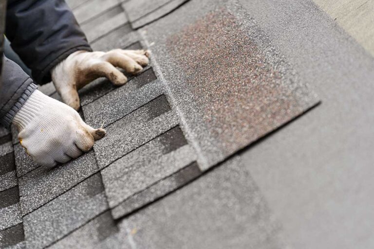 A person inspecting a residential roof with visible shingles and tree branches nearby.